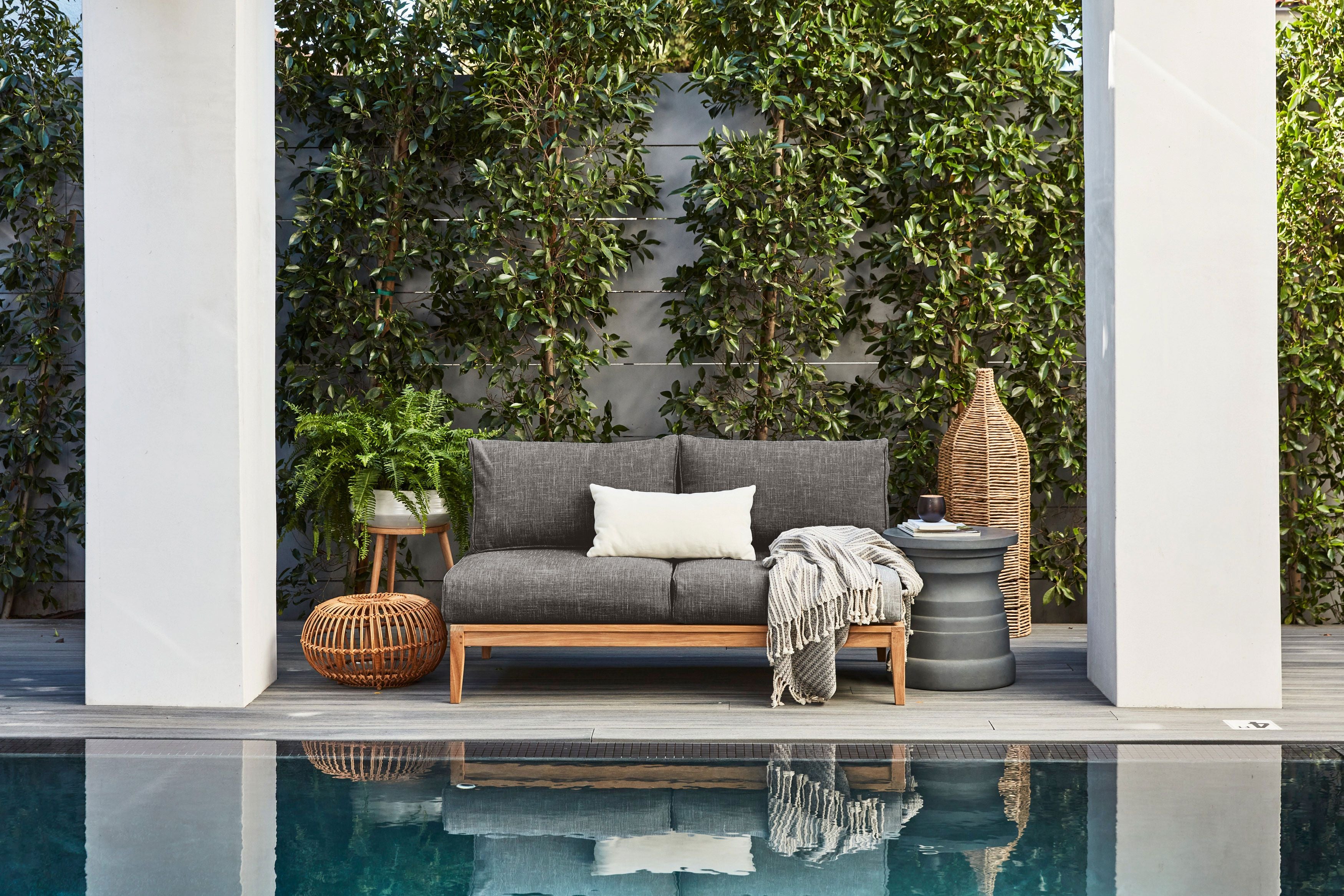 The outdoor teak armless loveseat poolside with pebble gray fabric and our gray tiered side table.