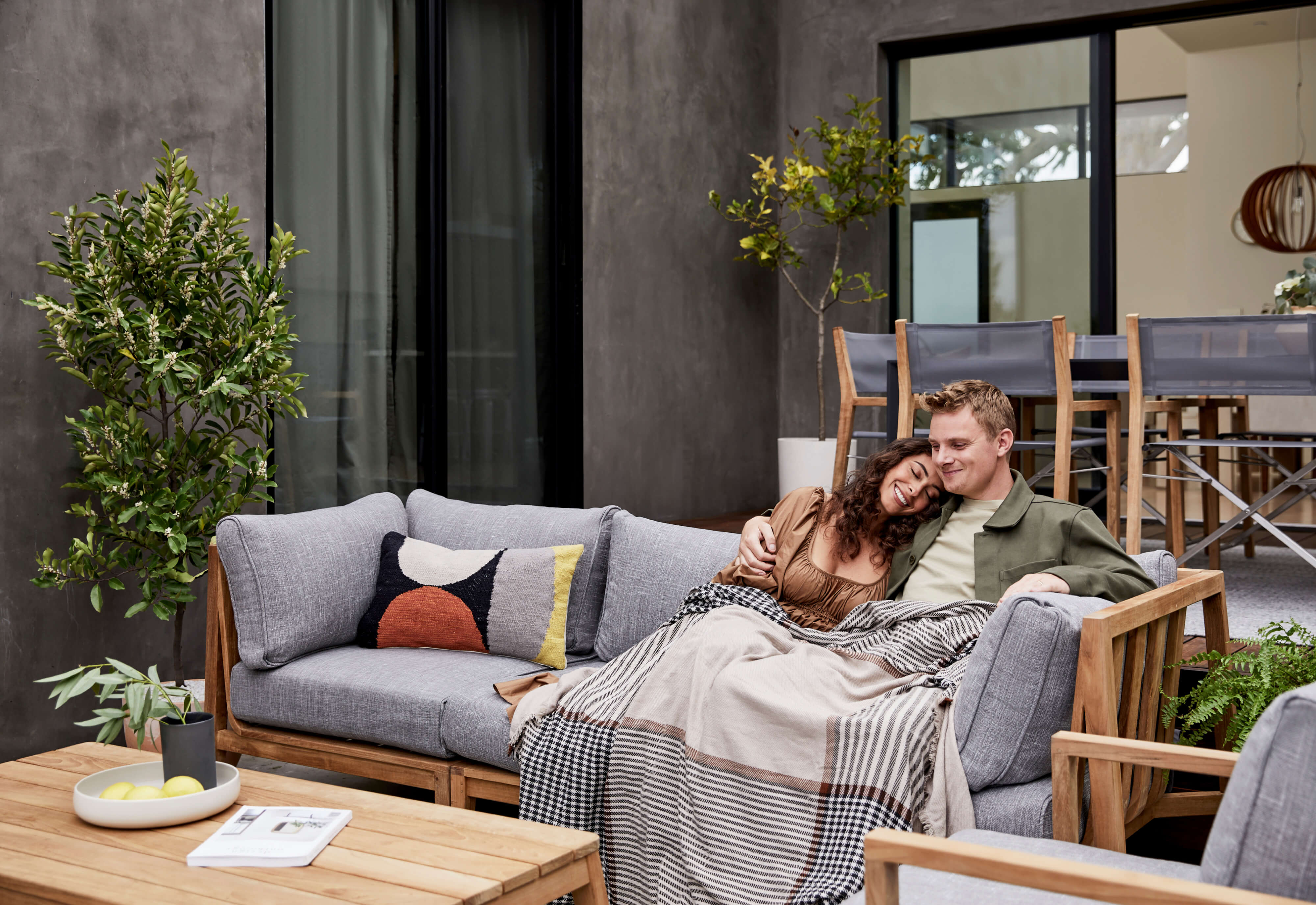 A couple sitting on the FSC Certified teak outdoor sofa wrapped in a striped plaid outdoor bug shield blanket.