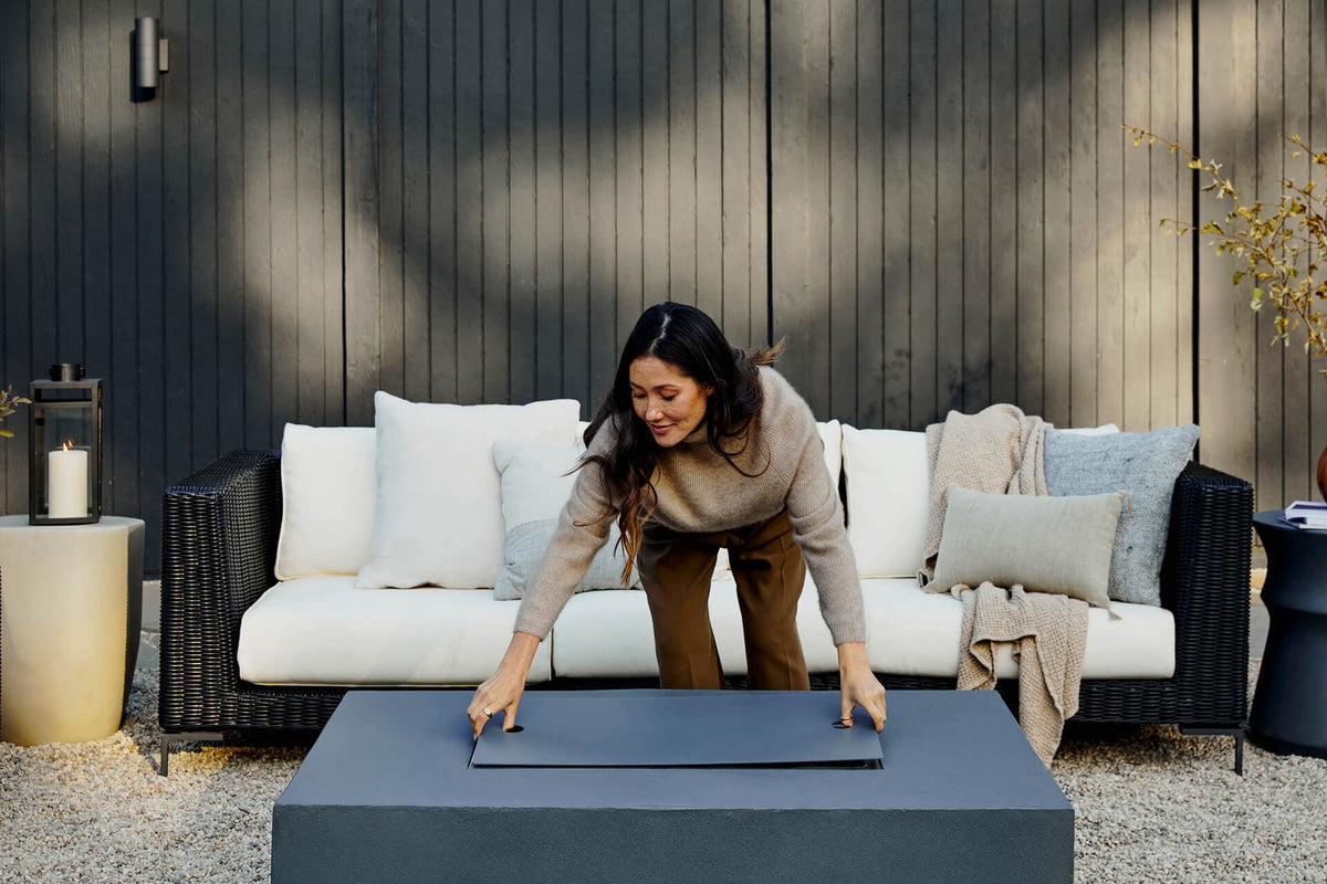 Woman placing a black metal burner cover lid onto a rectangular fire pit coffee table.