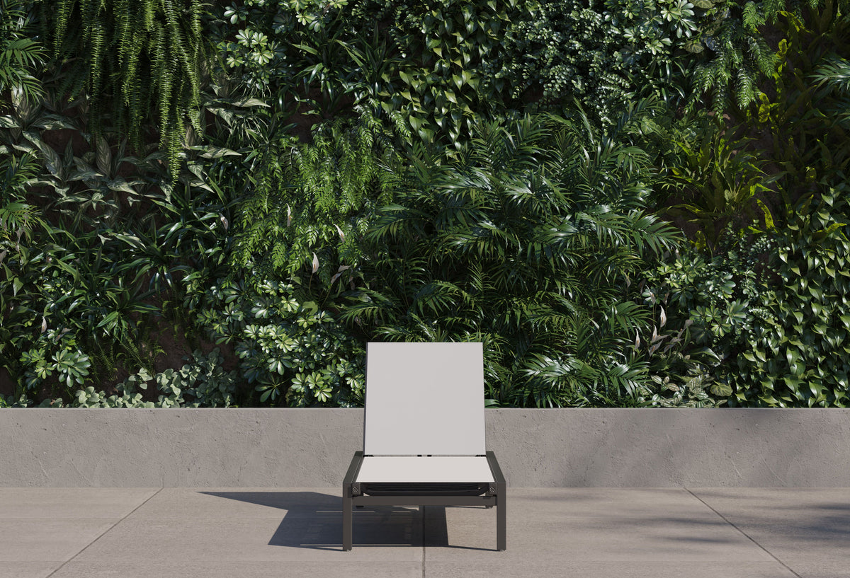 A charcoal aluminum Infinity chaise lounge with a light sling on a paved terrace in front of dense green planting.