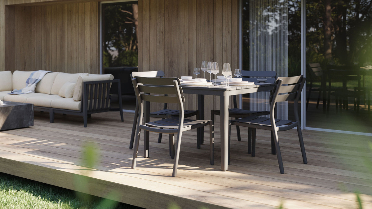 A square charcoal aluminum outdoor dining table set with four chairs, stemmed glasses, and plates, with a cushioned sofa nearby.