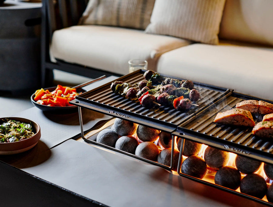 Vegetable skewers and salmon grilling on dual cast-iron cooktop grates over a lit fire pit table.