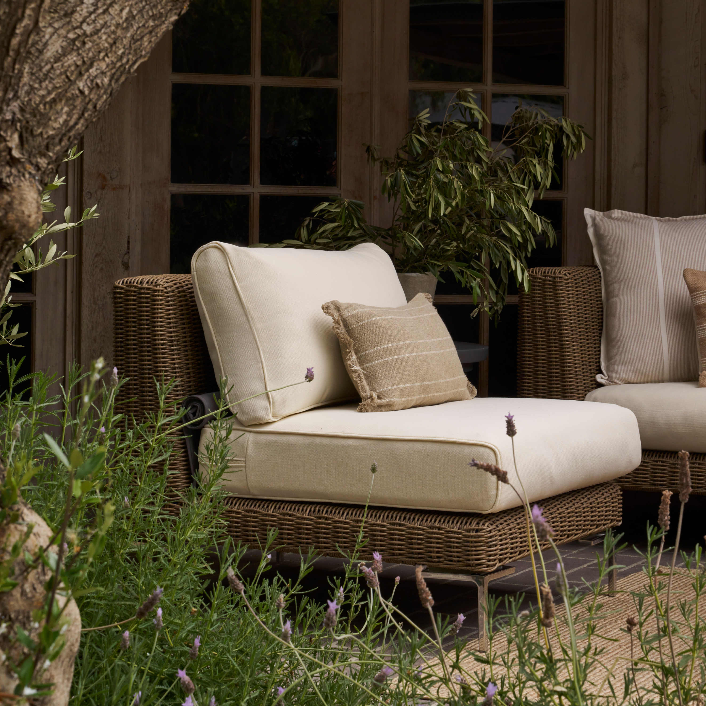 Brown wicker armless outdoor chair with cream cushions in a garden setting