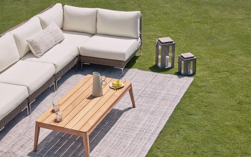 Outer teak outdoor coffee table with sectional sofa set on patio rug in modern backyard lounge setting