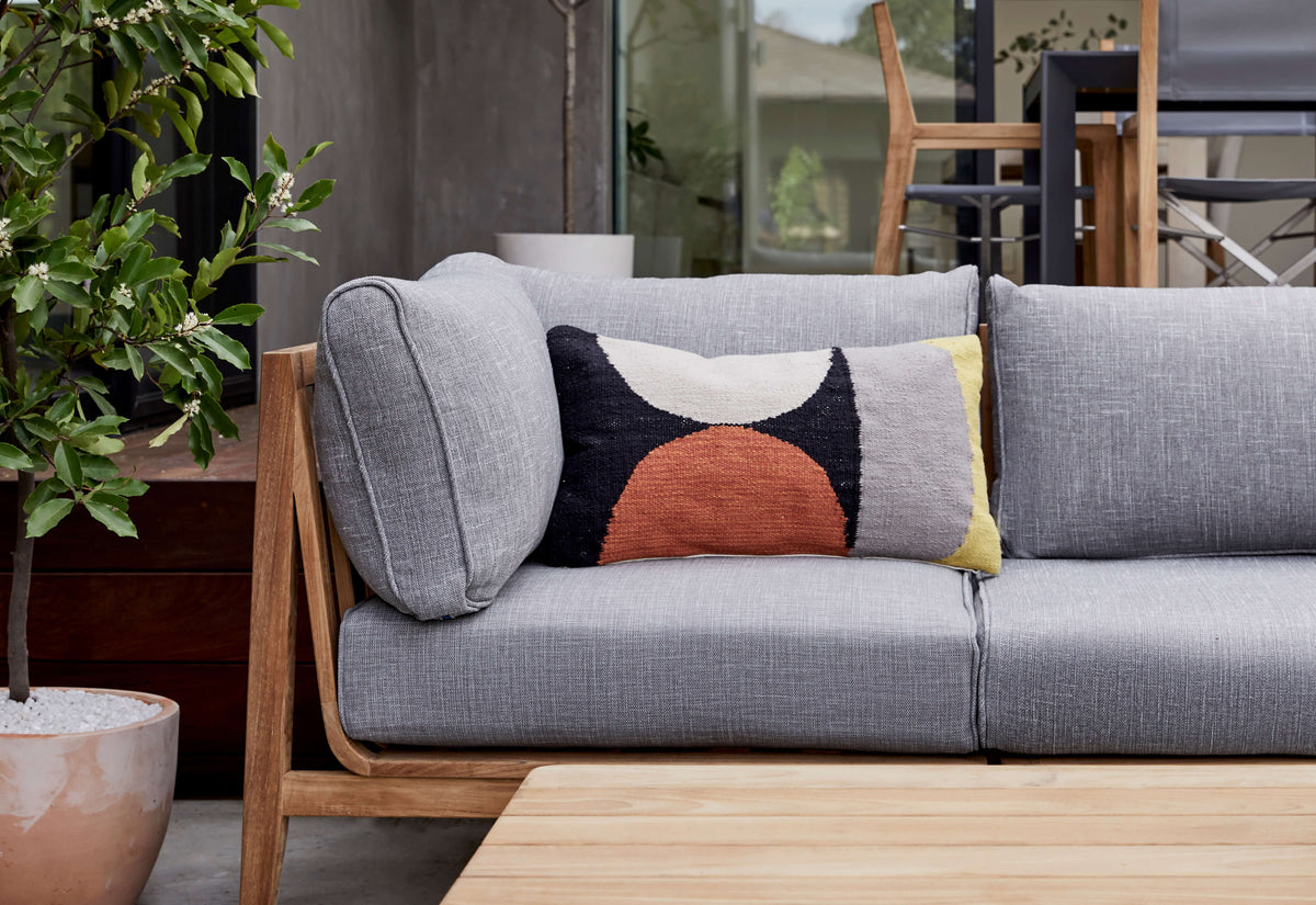 Close view of a teak outdoor sofa frame and armrest, gray seat and back cushions, and a geometric accent pillow.